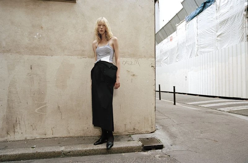 Fashion shot of a blond female model in a grey top and black skirt, posing against a textured beige wall in an urban environment.