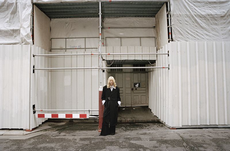 Woman in dark coat stands outside of a metal building; she has blonde hair, a neutral expression, and wears dark clothing and a white shirt.
