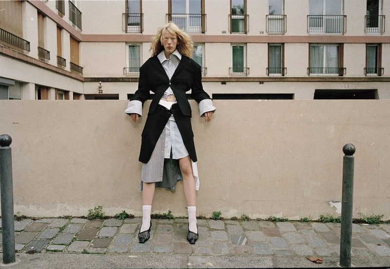 Fashionable woman in layered black coat and striped skirt poses against a concrete wall in an urban setting with building in the background.