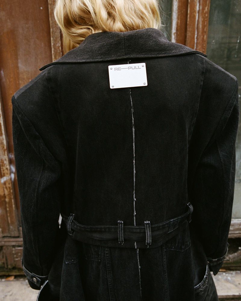 Back view of a person wearing a black trench coat with a gray collar, standing in front of an aged door. The coat has a belt at the waist.