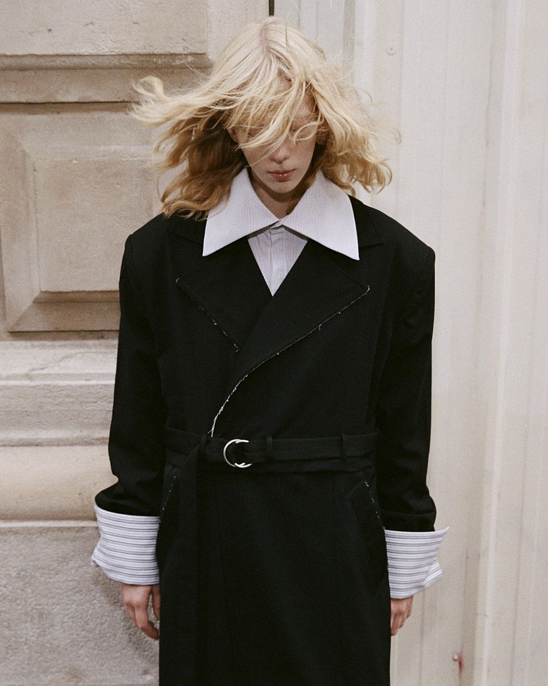 A person with blonde hair wears a black trench coat with an oversized white collar and striped cuffs against a neutral architectural backdrop.