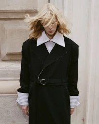 Model with blowing blonde hair wearing an oversized black coat and white collared shirt against a stone architectural background.