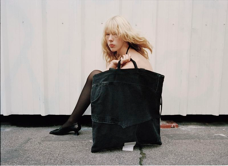 Blonde model sitting on the ground holding a large black denim tote bag in front of a white corrugated wall.