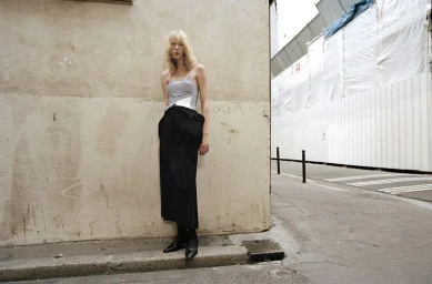 Model standing against a weathered concrete wall wearing a grey top and long black skirt on an urban street.