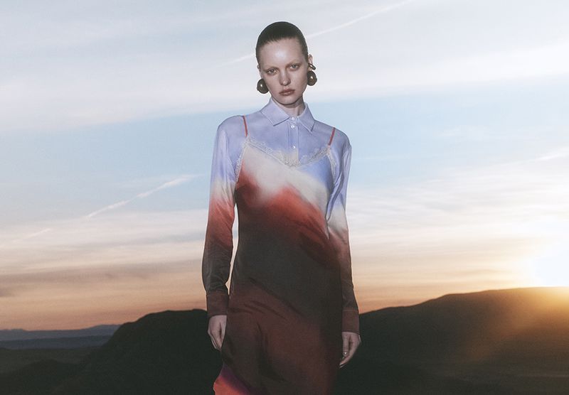 Model wearing a gradient slip dress over a collared shirt against a sunset sky and mountain landscape