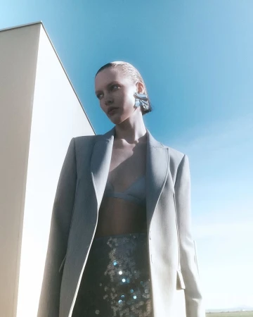 Woman wearing a blazer and earrings against a blue sky, in a fashion shot, with an architectural element and field in the background.