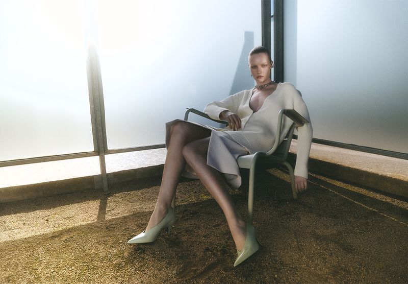 Model seated in an armchair wearing a light-coloured V-neck dress and heels on a balcony floor against a bright frosted glass backdrop