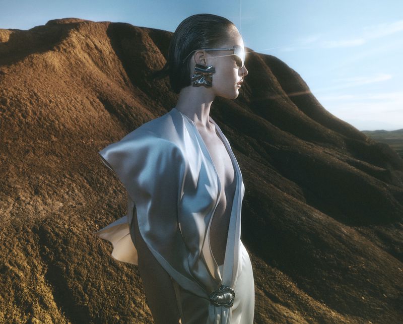 Fashion shot of a woman in a shimmering dress and sunglasses posing in a desert landscape with sandy dunes and clear sky.