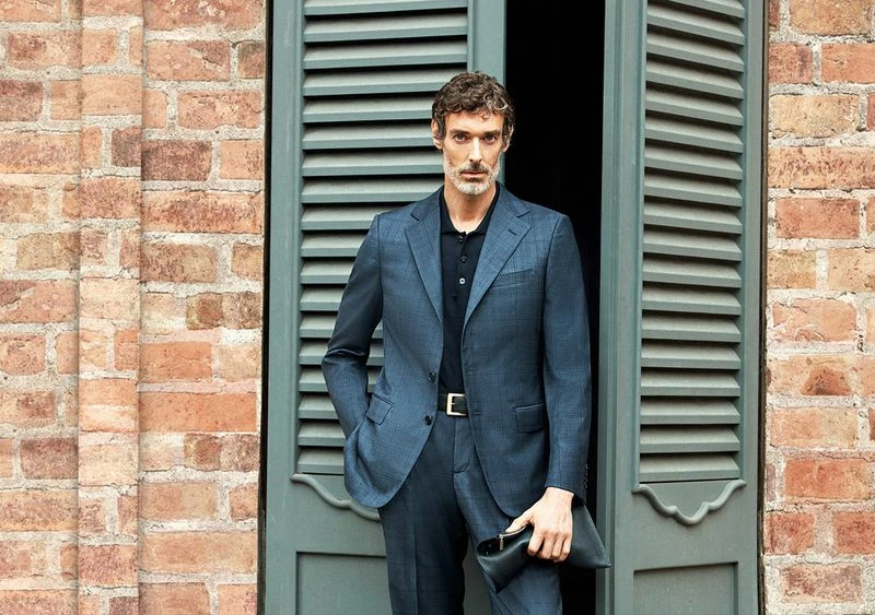 An adult man in a blue suit stands against a brick wall and shuttered window, holding a black clutch. He poses for a fashion shot.