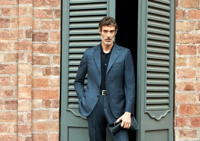 An adult man in a blue suit stands against a brick wall and shuttered window, holding a black clutch. He poses for a fashion shot.