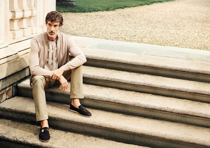 An adult man is sitting on outdoor stone steps, wearing a cardigan, tee shirt, pants, and loafers in a casual outdoor fashion portrait.