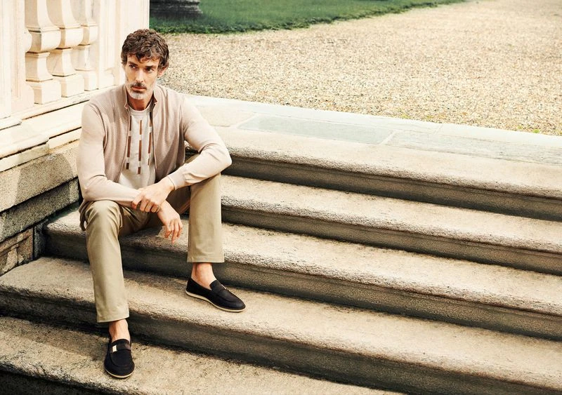 An adult man is sitting on outdoor stone steps, wearing a cardigan, tee shirt, pants, and loafers in a casual outdoor fashion portrait.