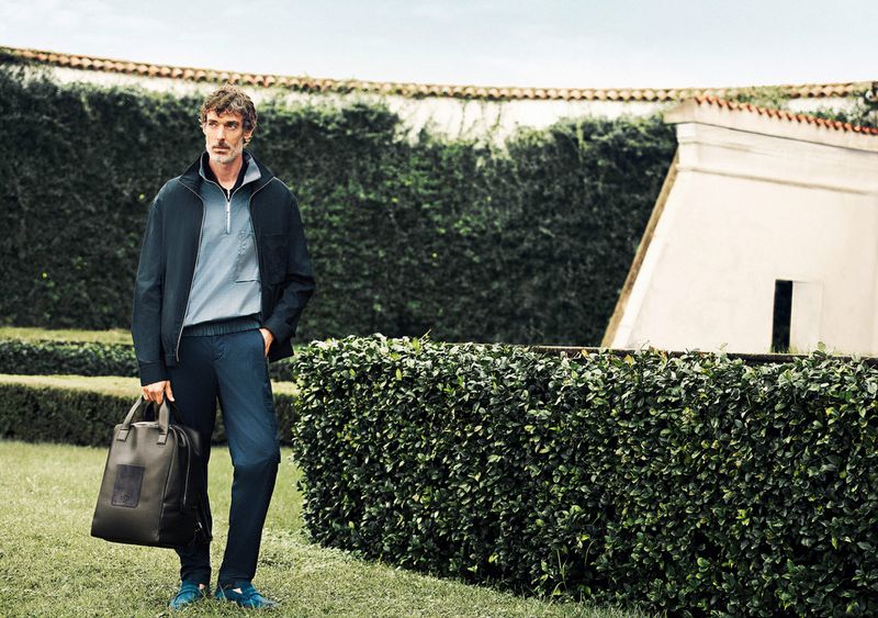 Model Richard Biedul in a dark jacket holding a leather bag standing outdoors near neatly trimmed hedges for Zilli Spring 25.