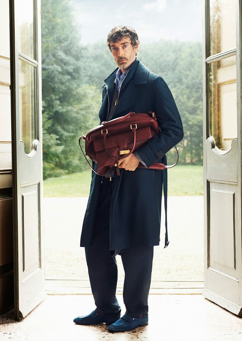 Adult male standing in doorway wearing navy overcoat, navy pants, and blue shoes holding a burgundy leather bag on a rural property.