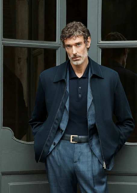Portrait of a mature adult man in dark blue jacket and patterned pants, leaning against a gray window frame, looking at the camera.