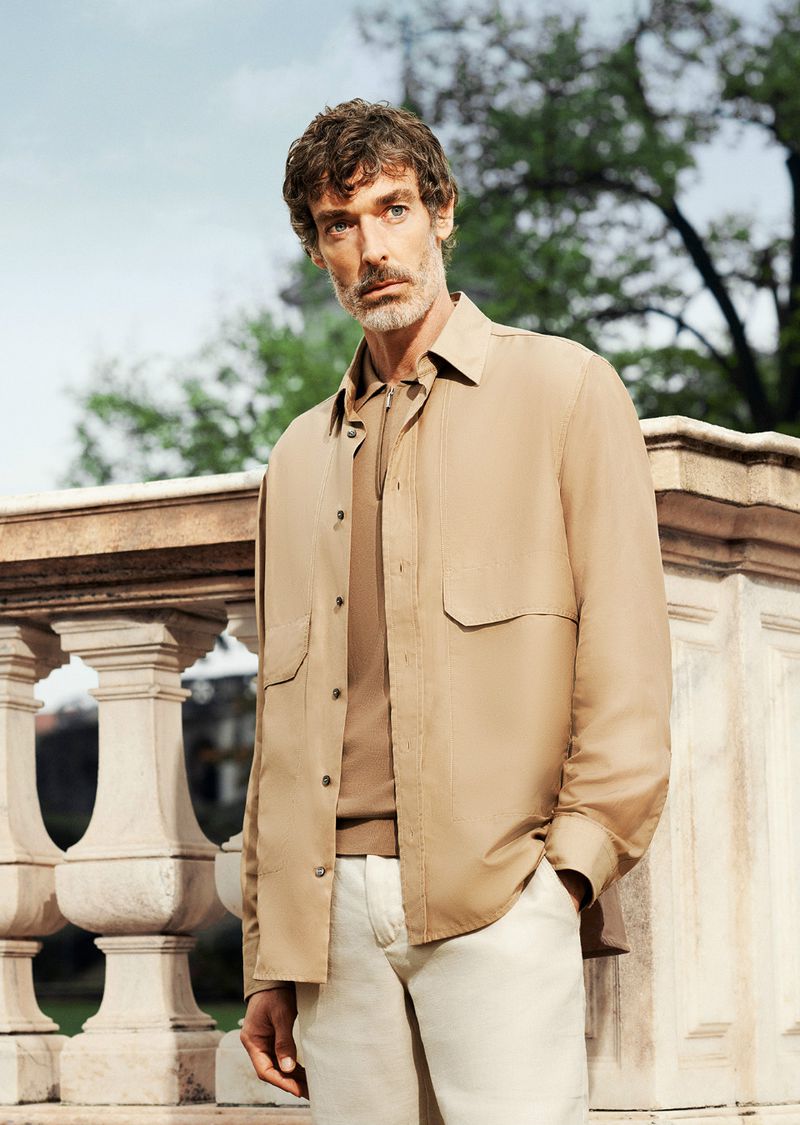 A man in a light brown shirt and white pants poses outside against a light sky and trees, next to a decorative stone railing.