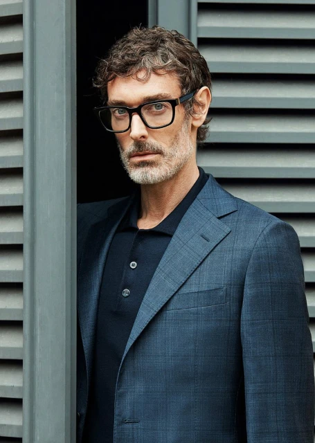 A fashionable adult man wearing a blue blazer and glasses poses next to a window with grey shutters for an indoor portrait.