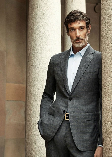 Portrait of a man in a gray suit standing between stone columns, looking at the camera with a neutral expression.