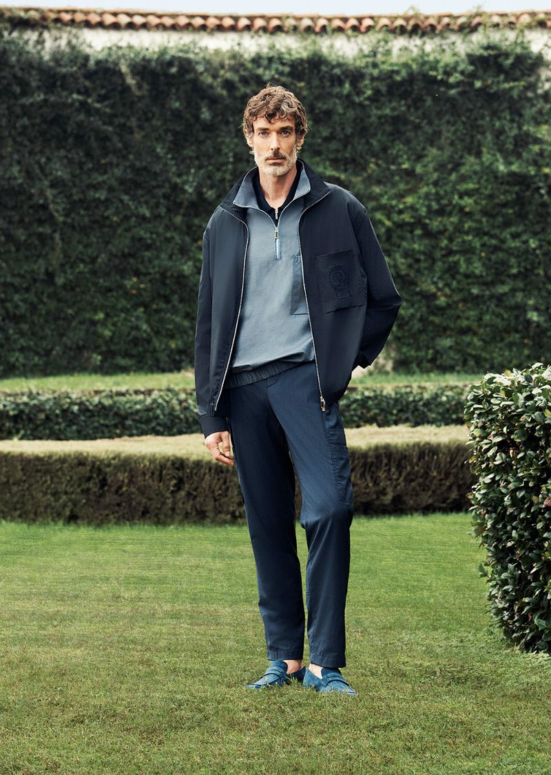 A man in navy jacket and blue pants stands on grass in front of a green hedge, looking at the camera with a neutral expression.