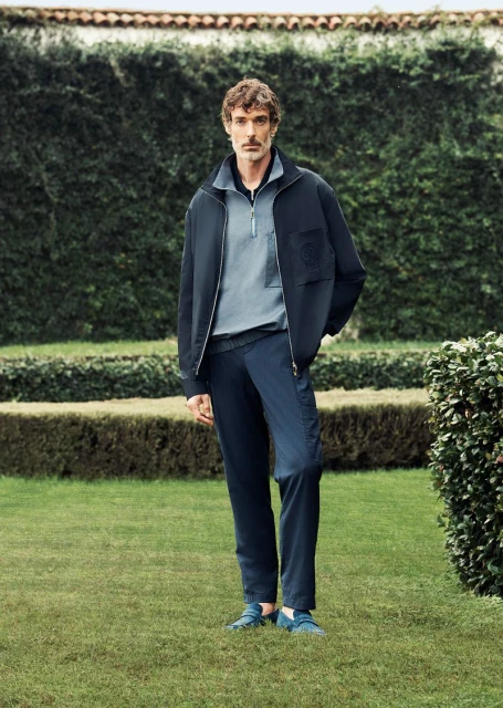 A man in navy jacket and blue pants stands on grass in front of a green hedge, looking at the camera with a neutral expression.
