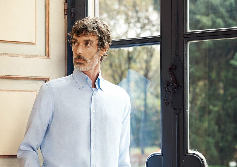 Middle-aged man in a light blue shirt poses indoors near a window, showcasing a contemplative gaze and stylish, casual fashion.
