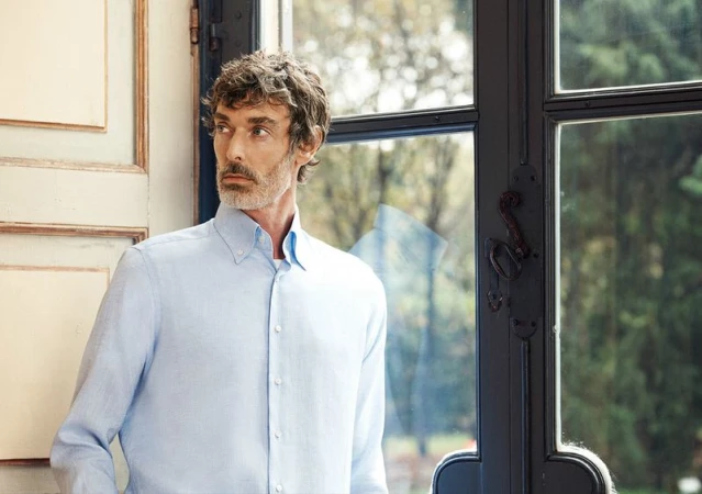 Middle-aged man in a light blue shirt poses indoors near a window, showcasing a contemplative gaze and stylish, casual fashion.