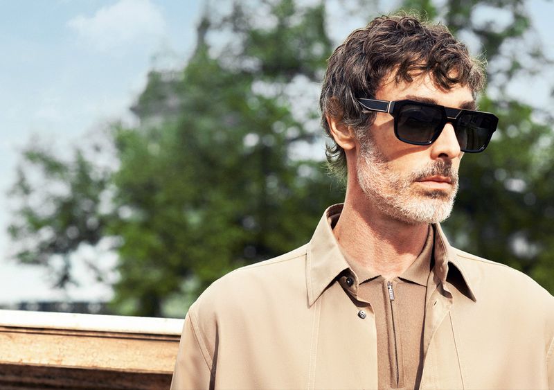Portrait of an adult man with dark sunglasses, short curly hair, and a tan jacket, posing outdoors with foliage and sky behind him.