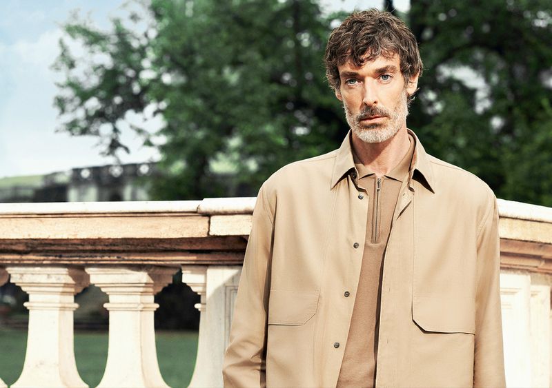 Portrait of an adult male wearing a beige jacket outdoors, featuring neutral tones and shallow depth of field against a green leafy background.