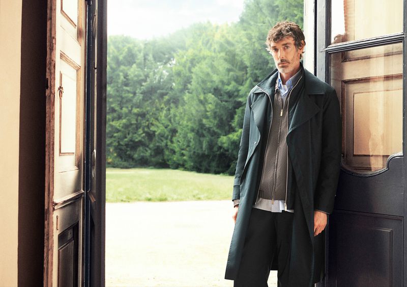 Adult man standing in an open doorway wearing a dark trench coat, sweater, and dress pants in front of a green, grassy landscape.