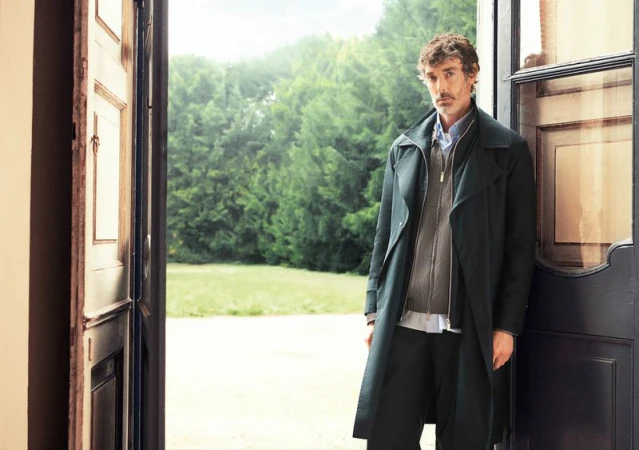 Adult man standing in an open doorway wearing a dark trench coat, sweater, and dress pants in front of a green, grassy landscape.