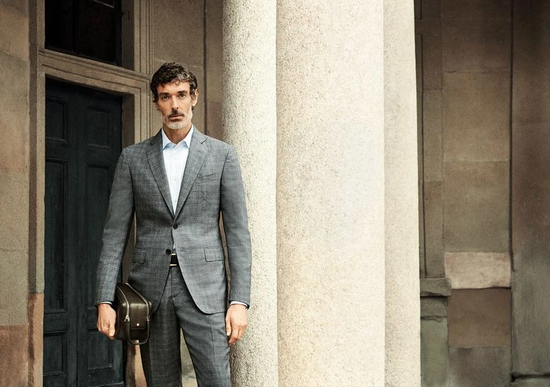 Well-dressed man in grey suit stands outside with briefcase near white stone pillars and an old door, looking at the camera.