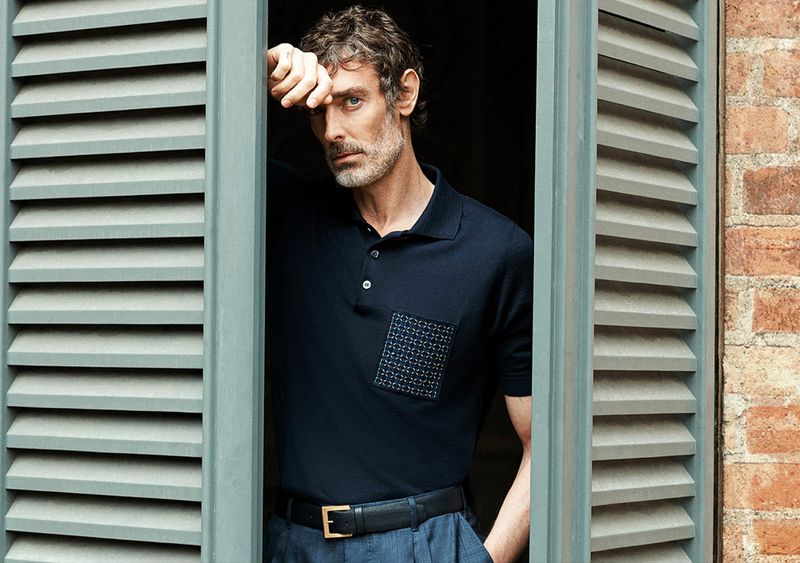 Adult male poses by a window with shutters, wearing a navy polo shirt and grey pants in an indoor setting with natural lighting.