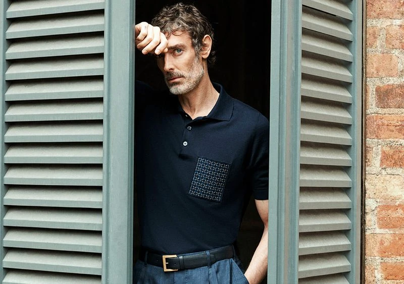 Adult male poses by a window with shutters, wearing a navy polo shirt and grey pants in an indoor setting with natural lighting.