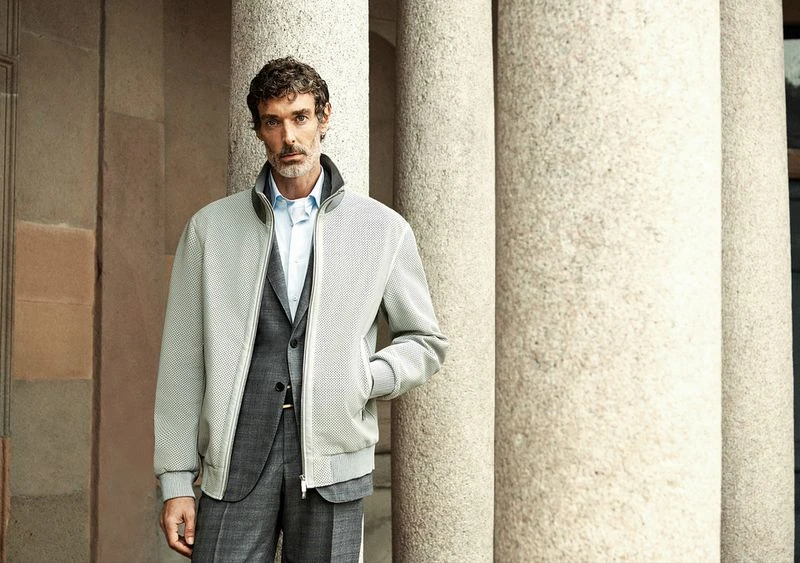 Adult male in a grey suit and light grey jacket stands in front of stone columns in an outdoor setting, captured in a medium shot.