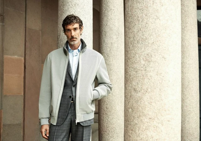 Adult male in a grey suit and light grey jacket stands in front of stone columns in an outdoor setting, captured in a medium shot.