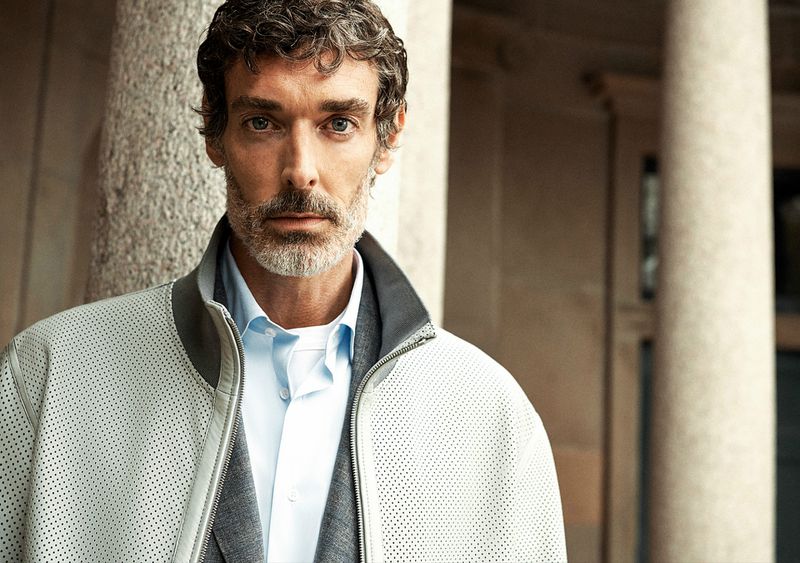 Portrait of an adult male with a light grey beard and jacket, wearing a light blue shirt, and standing in front of a building.