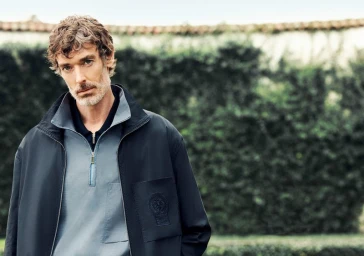 Model Richard Biedul wearing a dark jacket and blue zip polo shirt outdoors against a green hedge backdrop