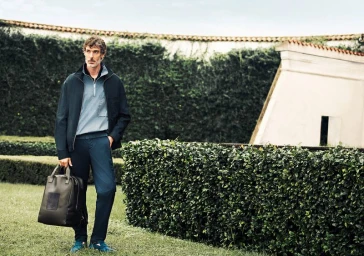 Model Richard Biedul in a dark jacket holding a leather bag standing outdoors near neatly trimmed hedges for Zilli Spring 25.