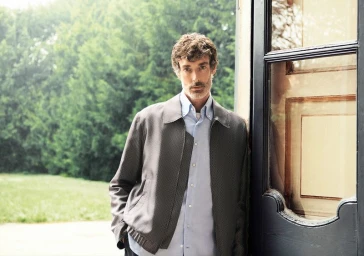 Man wearing a grey textured jacket and light blue shirt leaning against a dark door frame outdoors