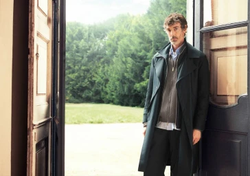 A man in a long dark coat standing in an open doorway with a lush green garden visible in the background.