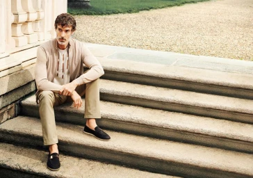 A man wearing a cream zip-up top, beige trousers, and loafers seated on outdoor stone steps.