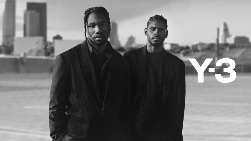 Two African-American men in dark coats posing in front of a blurred city skyline, Y-3 logo visible on the right, black and white.