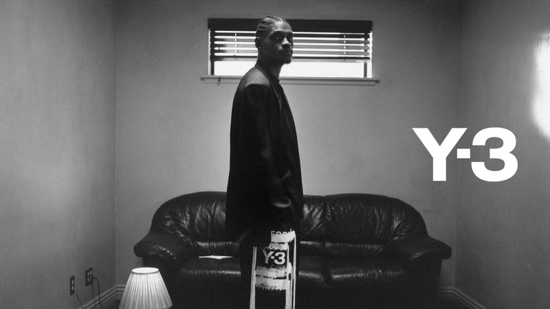 Black and white portrait of an adult male in a dark suit standing indoors next to a leather couch in front of a white wall.