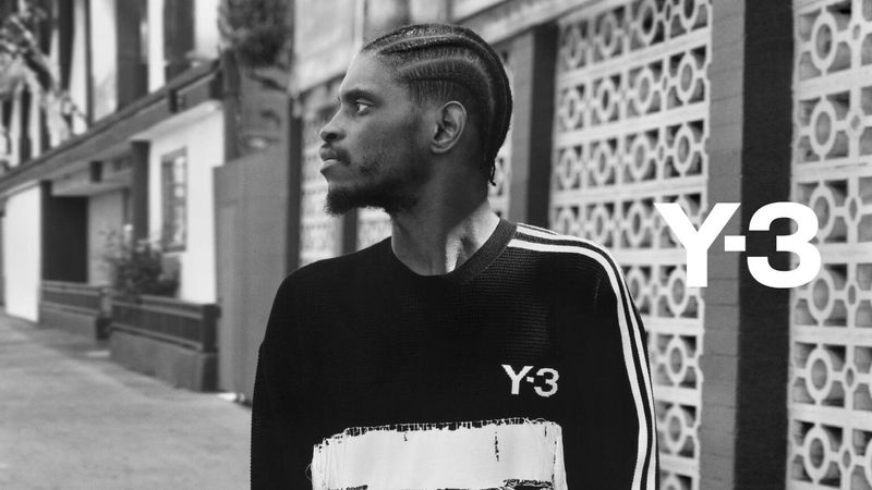 Monochrome portrait of a Black man with braided hair wearing a Y-3 sweater standing on a street in front of a decorative wall.