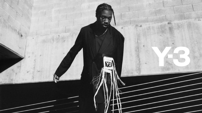 Fashion shot of a Black male model wearing a black suit and an accessory with white fringe against a modern concrete building with the Y-3 logo.