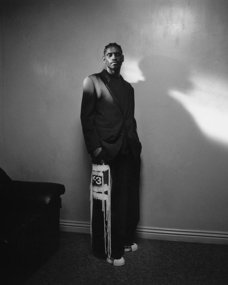 Black and white portrait of a male model in a dark suit holding a branded bag, standing in front of a neutral wall with dramatic lighting.