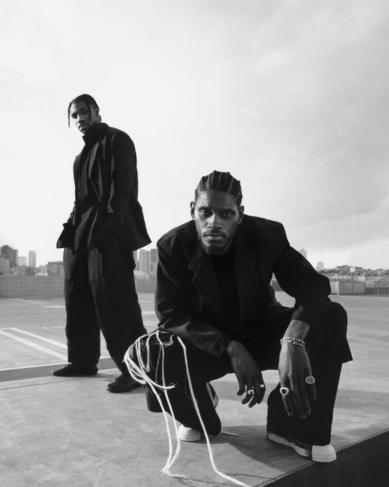 Two men in dark jackets pose on a rooftop against a city skyline. One is crouching, while the other stands behind him in a monochrome outdoor shot.