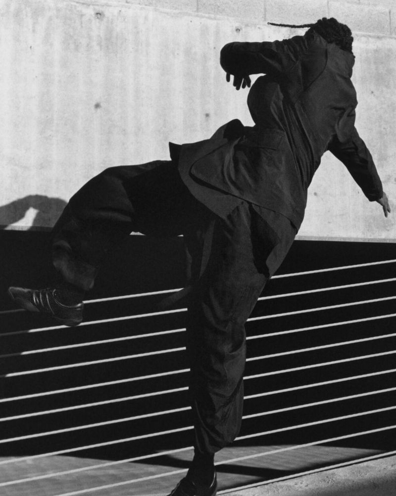 Monochrome action shot of a person in mid-air wearing a jacket, pants, and sneakers, against a textured wall with horizontal stripes.