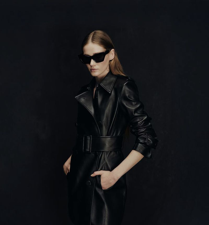 Portrait of a stylish woman wearing a black leather trench coat and sunglasses against a dark background in a fashion-forward pose.
