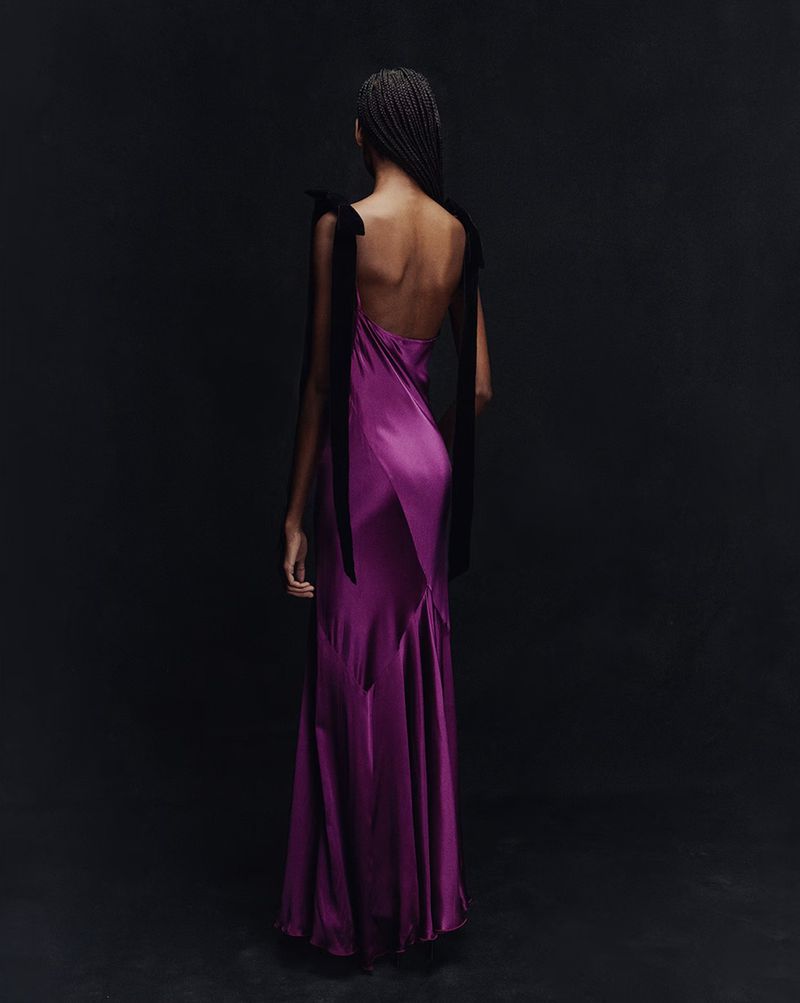 Back view of a slender adult female posing in a purple satin gown with black ribbons, set against a dark black backdrop in a studio.